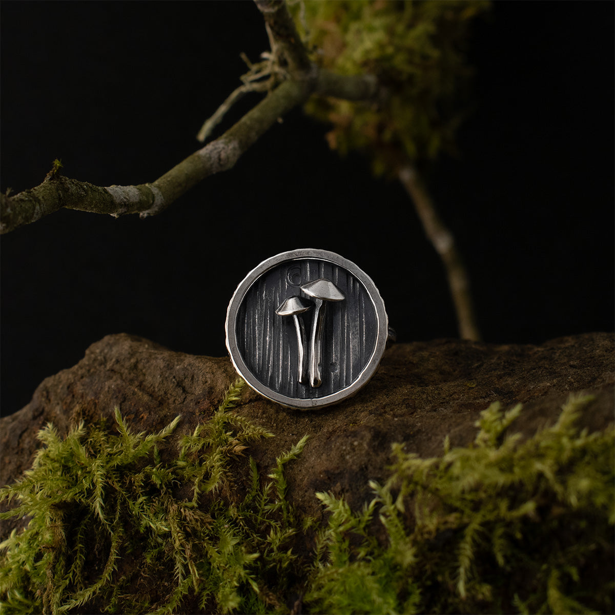 The Mycena Duo Shadow Box Ring is shown directly en face, its two sterling silver mushrooms surrounded by a darkly patinated bark background.