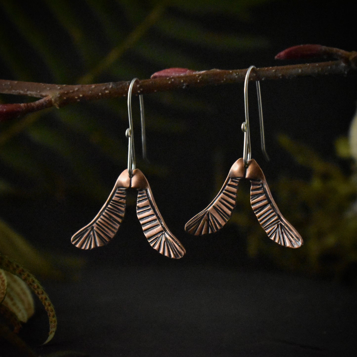 A pair of handmade copper dangle earrings, each stamped and textured to look like two maple seeds.