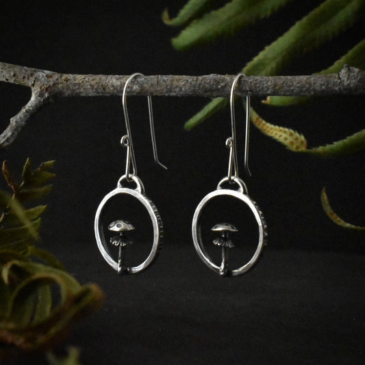 A pair of handmade sterling silver hoop earrings, each formed and textured to look like an amanita mushroom inside of a circle of tree bark.
