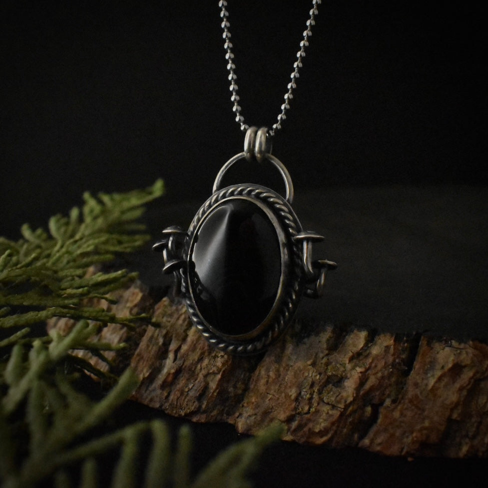 A handmade sterling silver pendant necklace, with a large setting of rich black onyx, surrounded by twisted wire and five formed mushroom elements, and hanging by a stainless steel ball chain.
