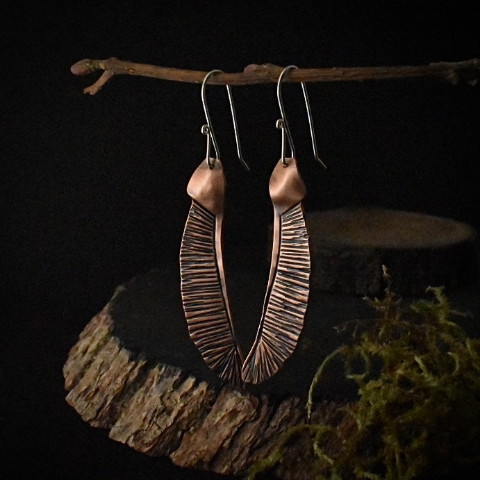 A pair of handmade copper dangle earrings, each stamped and textured to look like a maple seed.