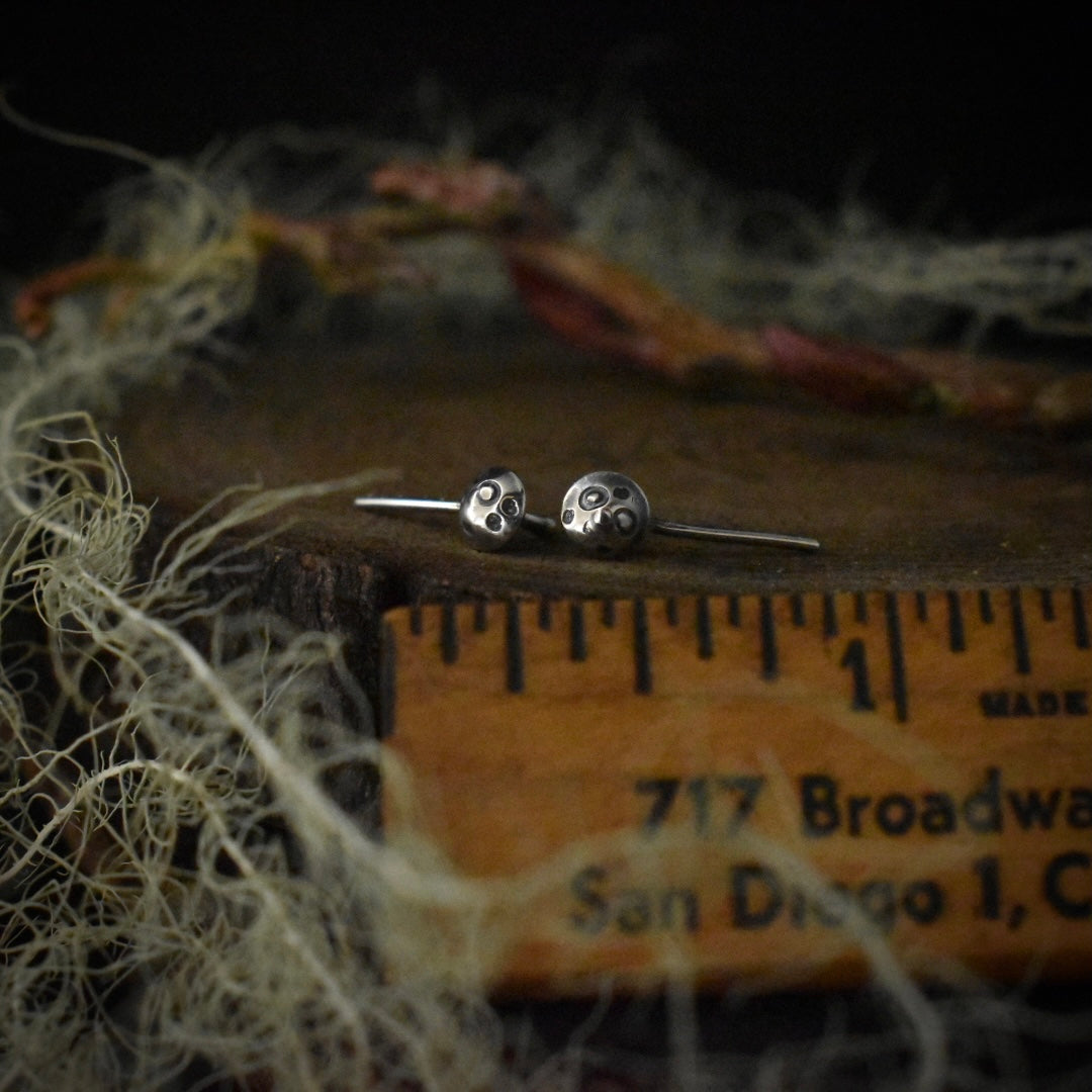 The Tiny Toadstool Stud Earrings with a ruler for scale, each mushroom cap measuring around 1/8 inch wide.