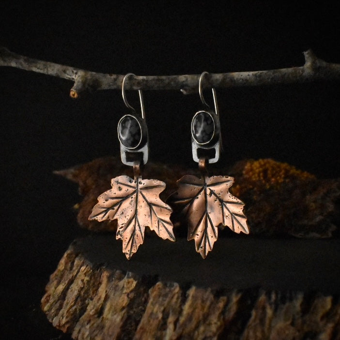A pair of handmade mixed metal dangle earrings, each with a textured copper maple leaf hanging from a setting of snowflake obsidian formed in sterling silver.
