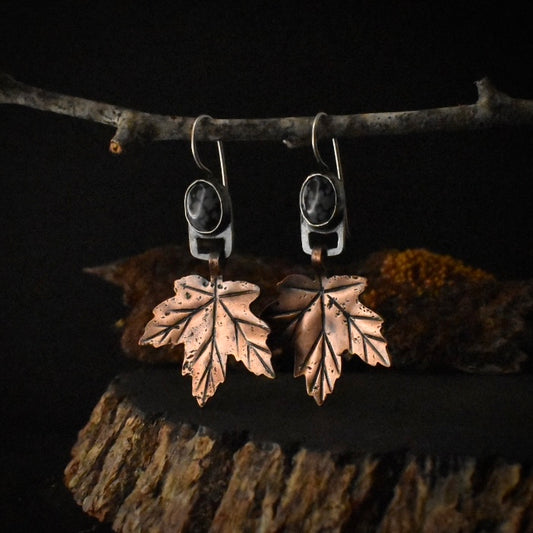 A pair of handmade mixed metal dangle earrings, each with a textured copper maple leaf hanging from a setting of snowflake obsidian formed in sterling silver.