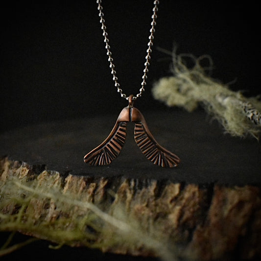 A handmade copper pendant necklace, stamped and textured to look like two maple seeds, hanging by a stainless steel ball chain.