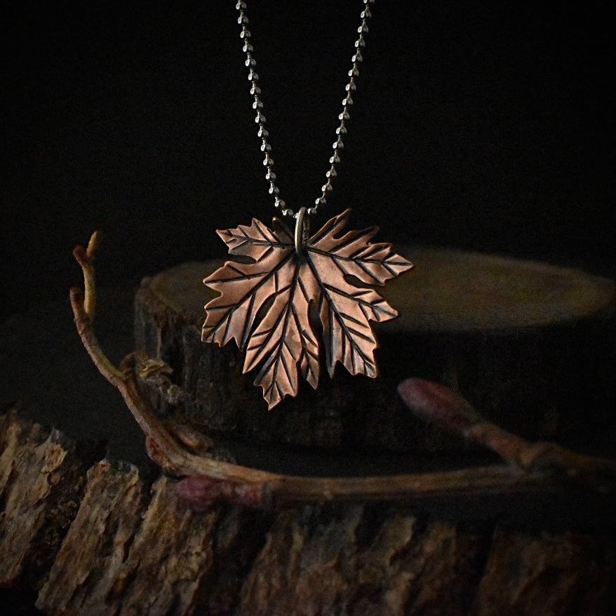 A handmade copper pendant necklace, stamped and textured to look like a maple leaf, hanging by a stainless steel ball chain.