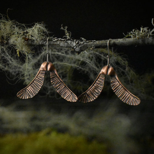 A pair of handmade copper dangle earrings, each stamped and textured to look like two maple seeds.
