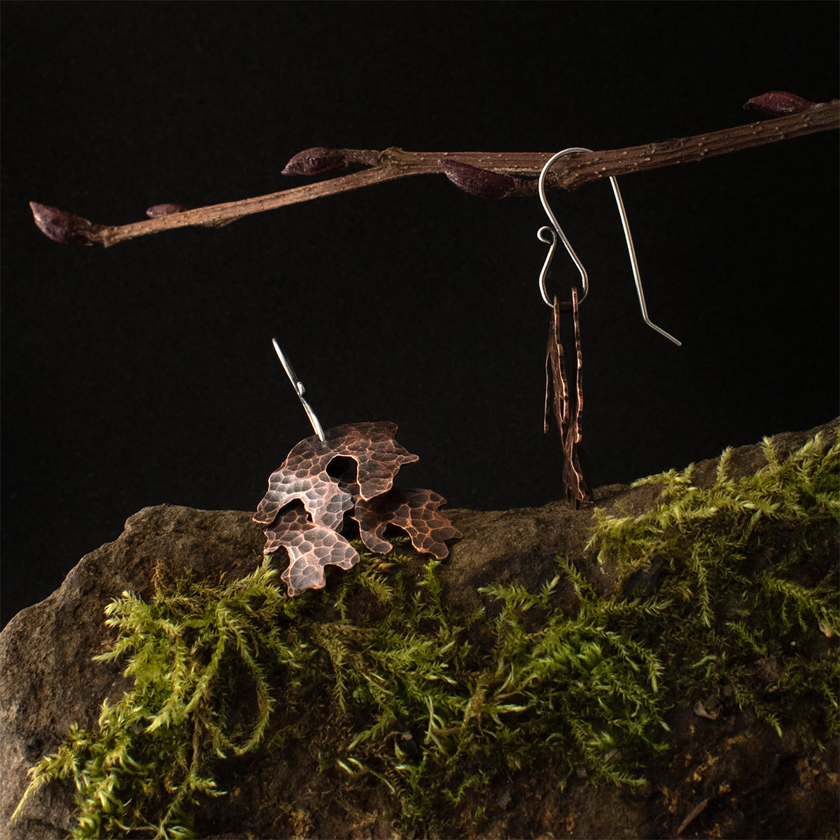 One of the Copper Layered Lichen Earrings is lying down, showing its richly patinated hammer marks, while the other earring dangles by its sterling silver ear wire, showing its two freely hanging layers of copper lichen.
