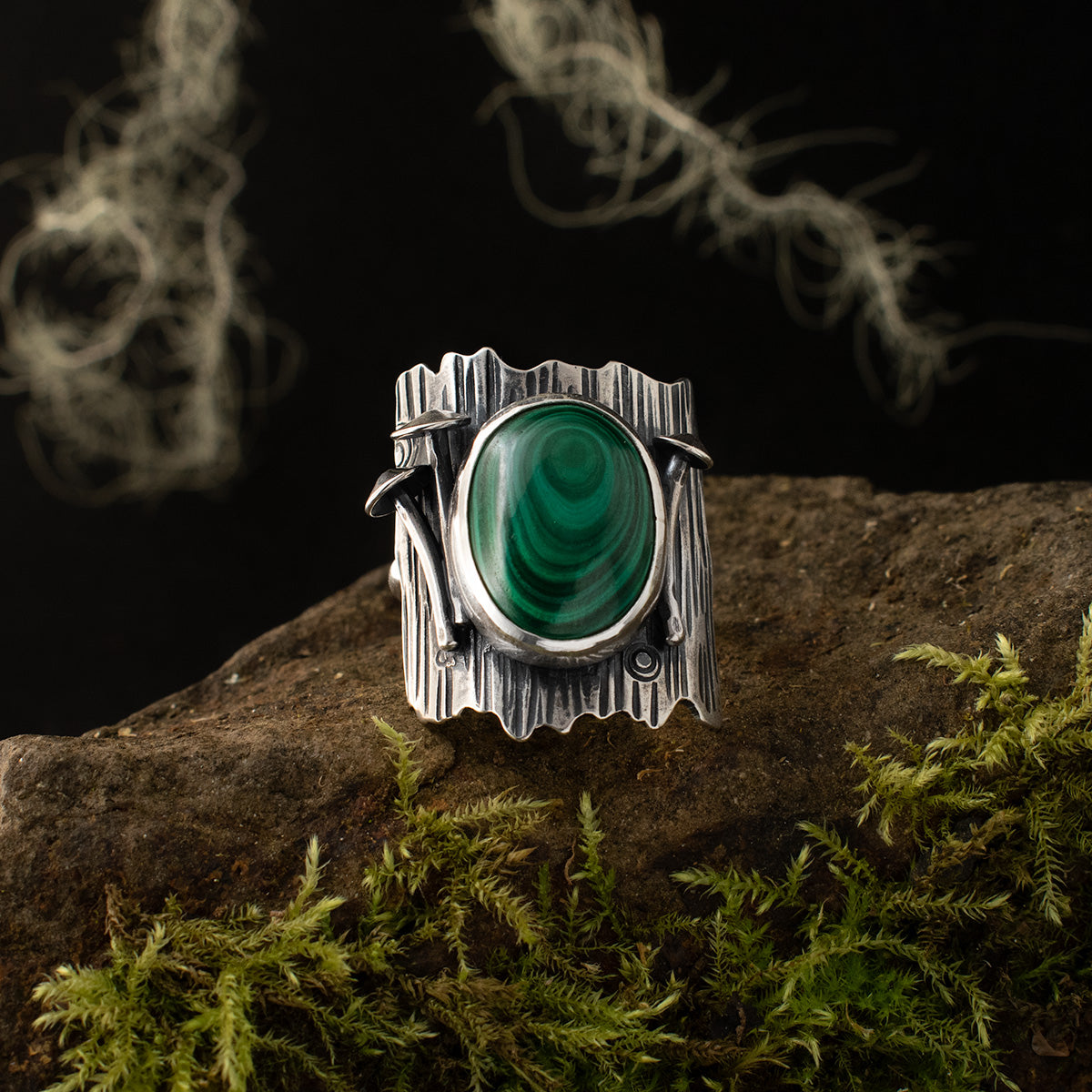 A handmade sterling silver statement ring, its large, bezel-set malachite, a spiral of green, is flanked by three miniature mushrooms and surrounded by sterling silver sheet metal, textured like a tree trunk with intricately stamped and patinated details.