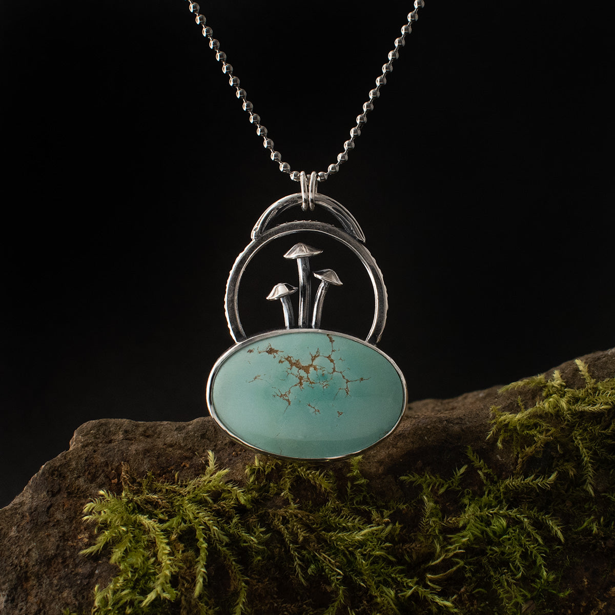 Three miniature mushrooms seem to spring up from this handmade sterling silver pendant’s oval cabochon of turquoise, webbed with tawny inclusions, as loops emerge around and above them.