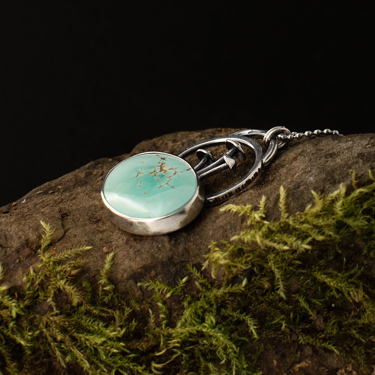 The Turquoise Fungal Loop Pendant lies down, showing its soft blue-green stone, its patch of sterling silver fungi, loops, and double bail.