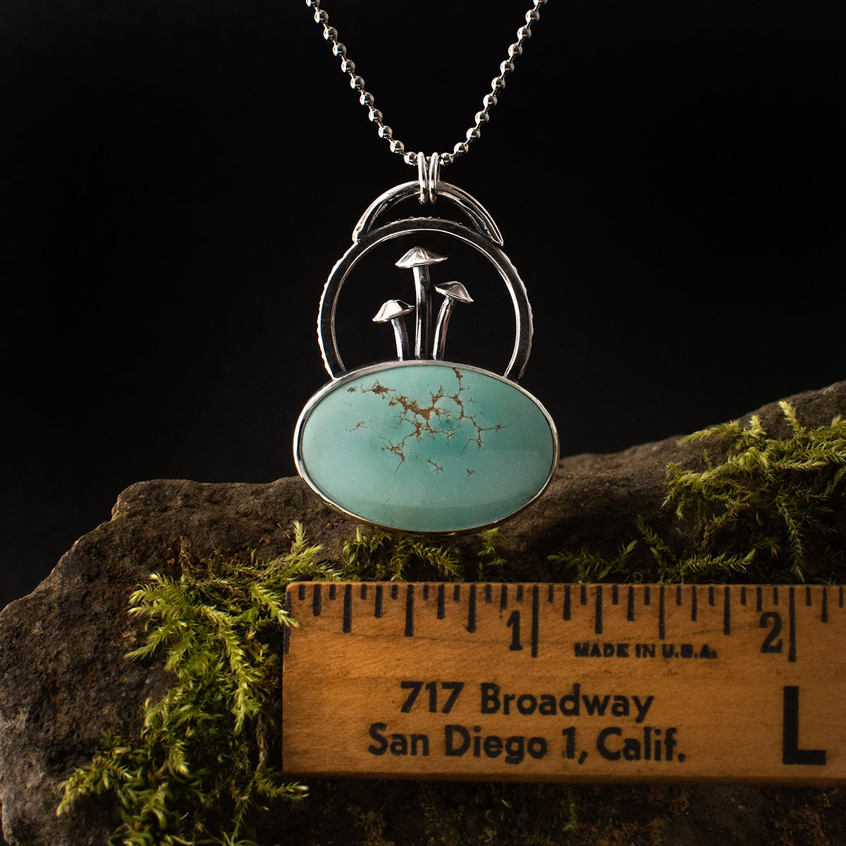 The Turquoise Fungal Loop Pendant with a ruler for scale, measuring around 1 and 1/4 inches wide from one end of the oval-shaped stone setting to the other.